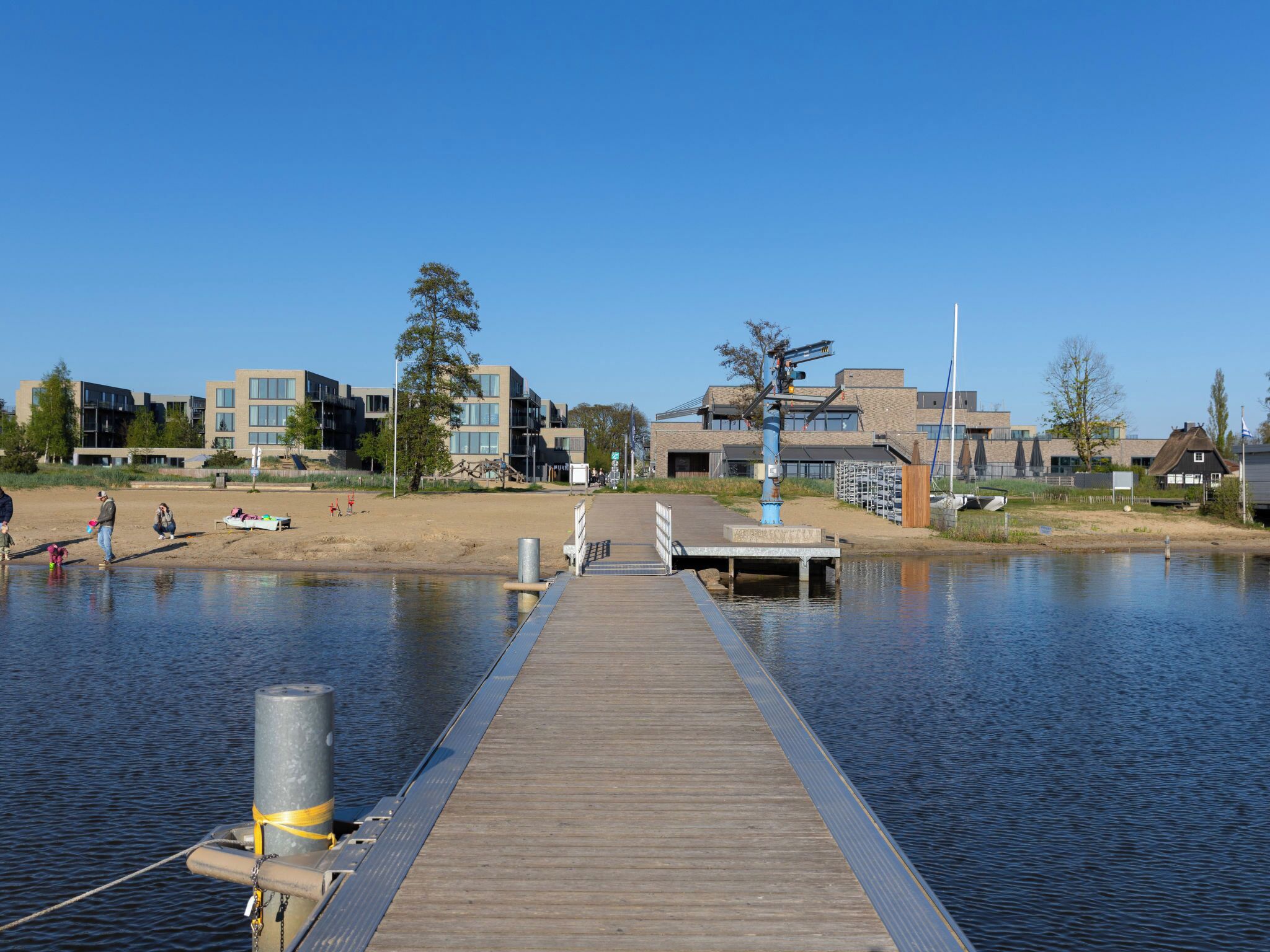 A wooden pier extending into calm water, with modern buildings and a sandy area in the background.