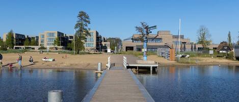 A wooden pier extending into calm water, with modern buildings and a sandy area in the background.