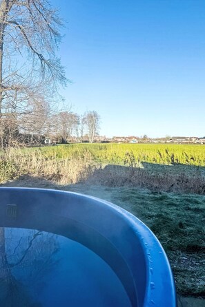 On voit clairement une piscine gonflable bleue, située dans un cadre rural, avec un champ et des maisons en arrière-plan.