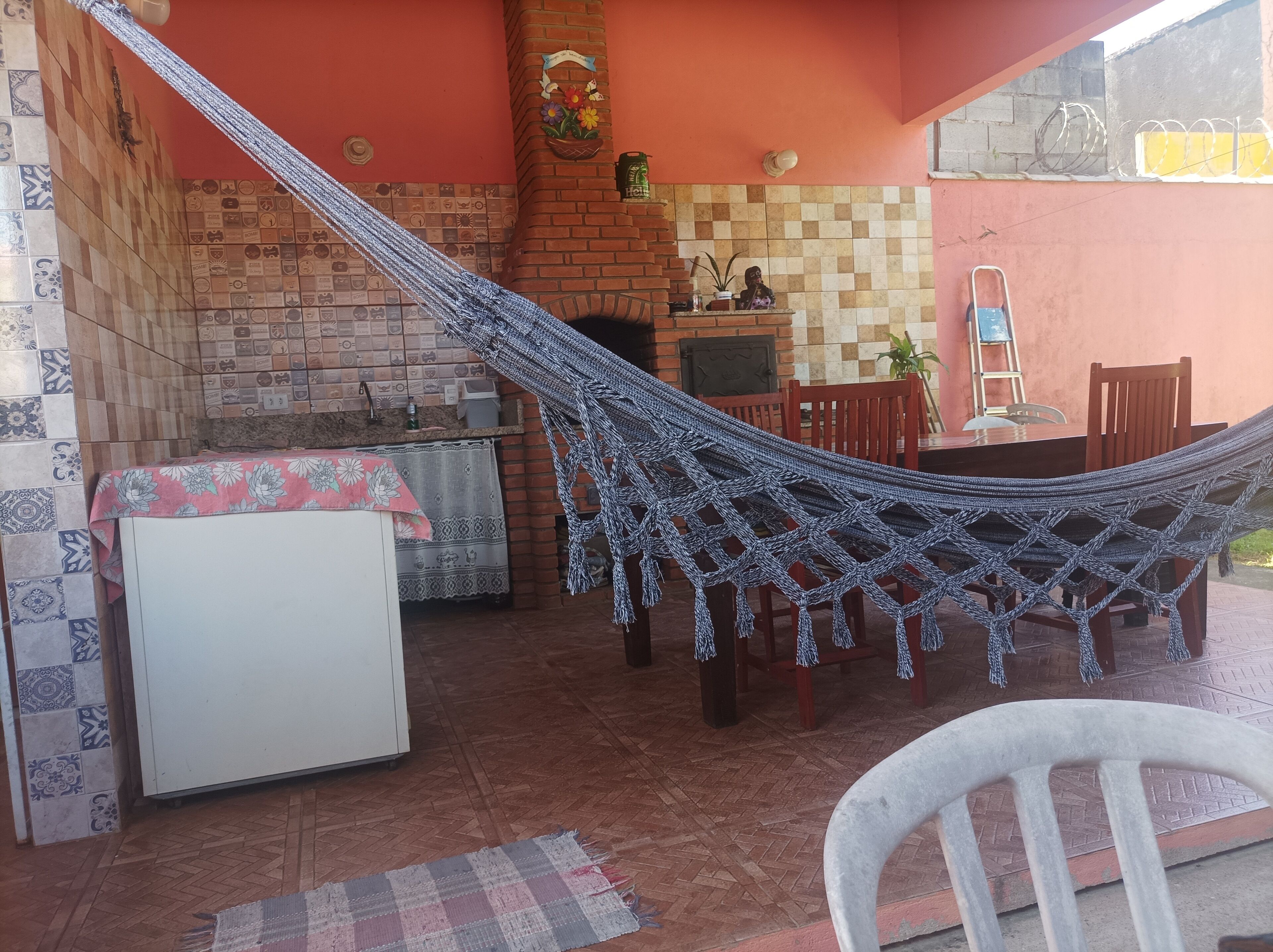 A hammock in a covered outdoor area with a brick wall, a small table, and a refrigerator.