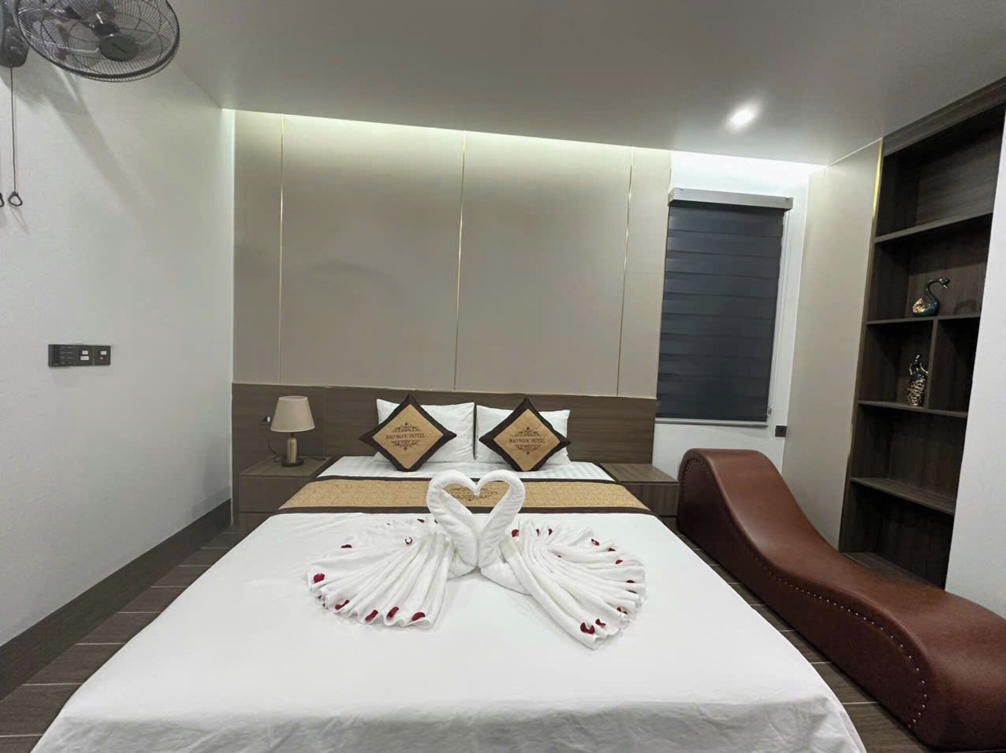 Una habitación de hotel con una cama bien hecha, un sillón de cuero marrón, una mesita de noche con una lámpara y un ventilador fijado a la pared.