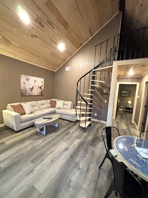 Un salon moderne avec un plafond en bois, un escalier en colimaçon, un canapé d’angle, une table basse et un coin repas avec une table en verre.