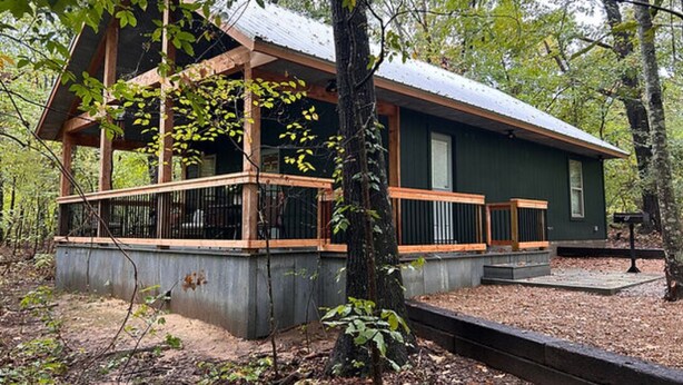 Eine Holzhütte mit überdachter Veranda, Betonfundament und Metalldach, umgeben von Bäumen.