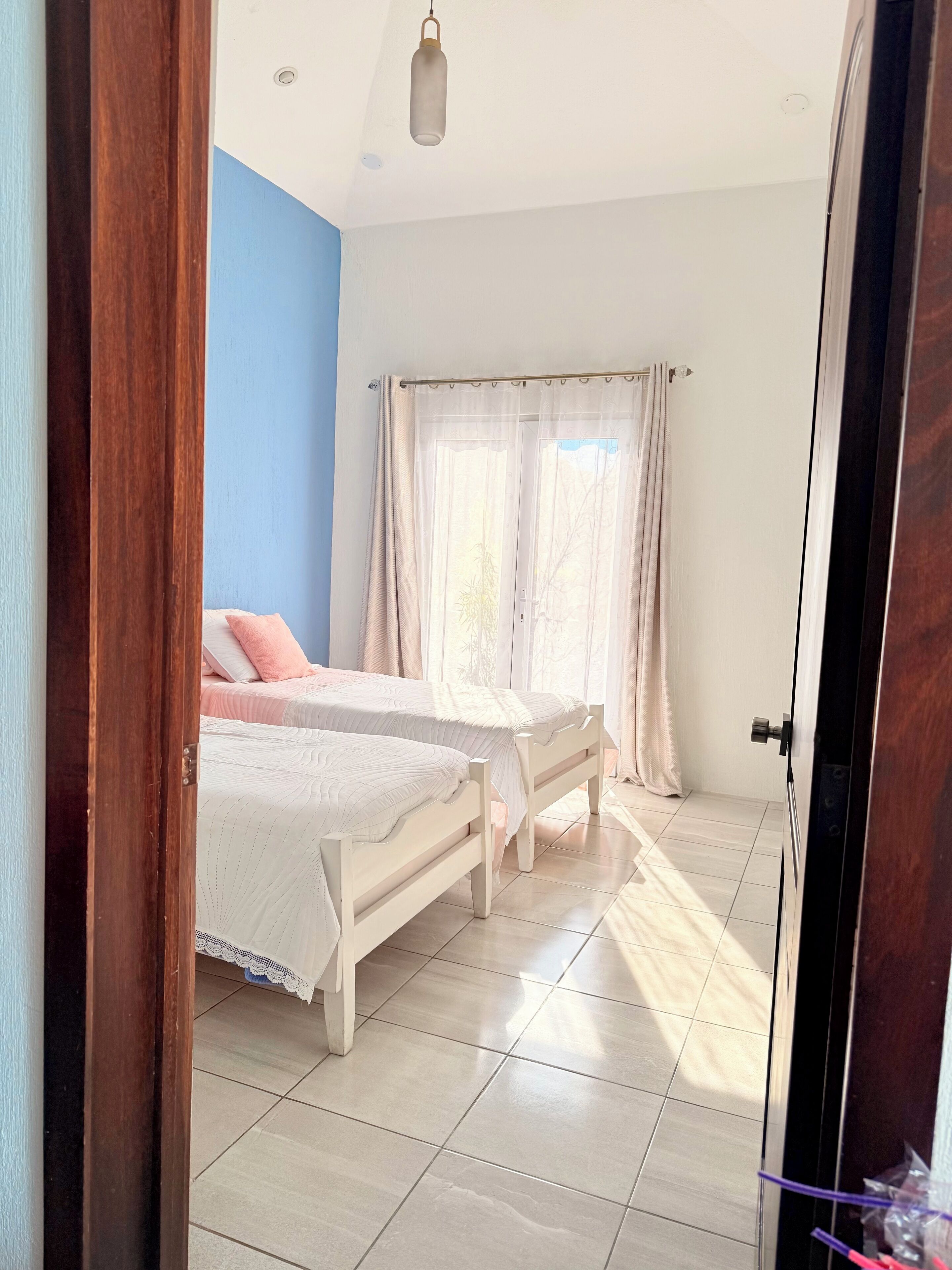Un dormitorio con dos camas, una ventana con cortinas translúcidas e iluminación en el techo.