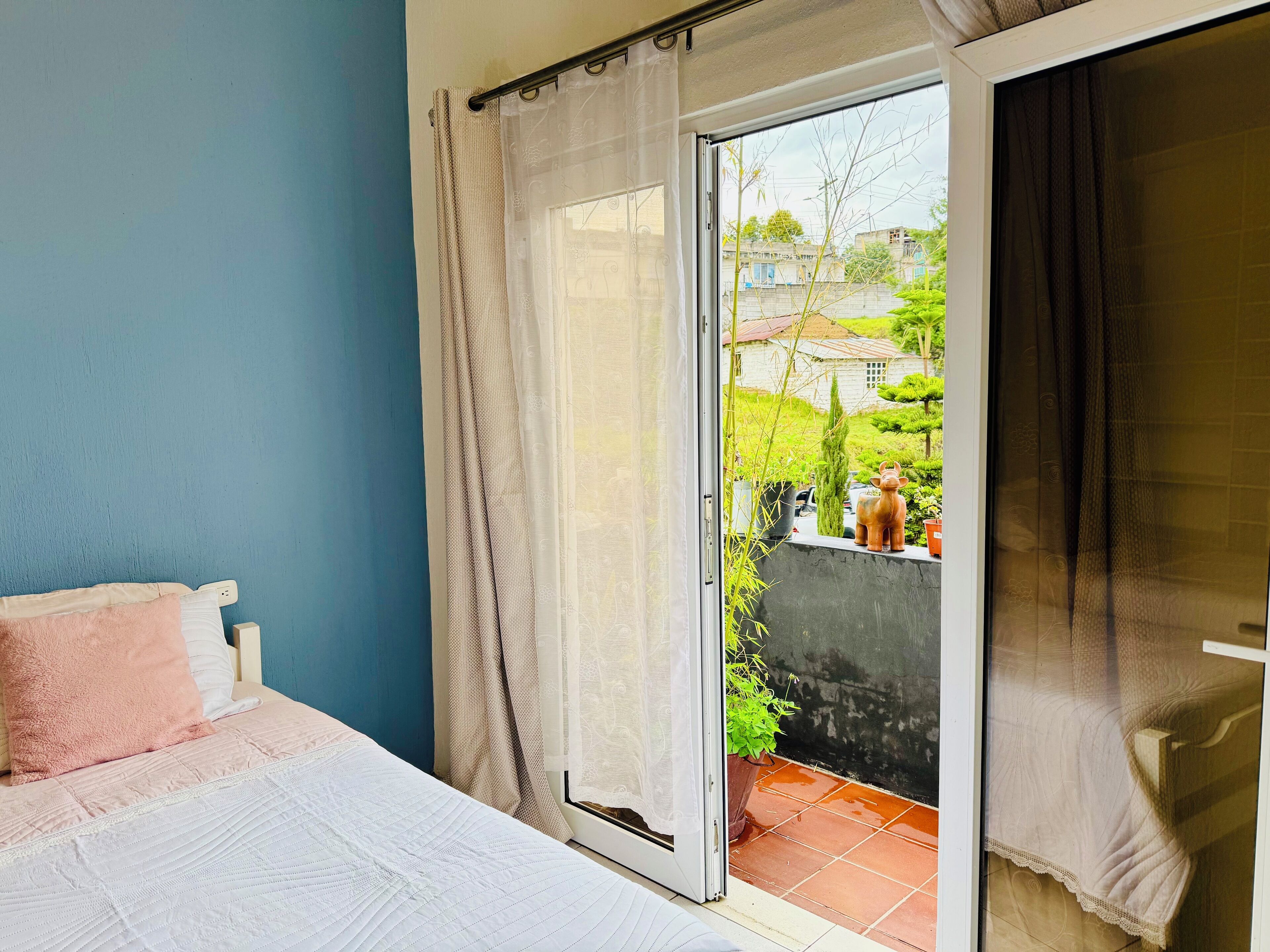 Un dormitorio con cama, ventana con cortinas y vistas a un jardín y edificios.