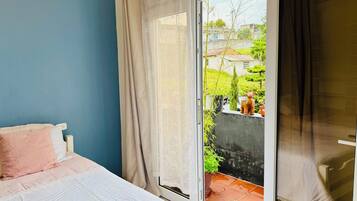 Un dormitorio con cama, ventana con cortinas y vistas a un jardín y edificios.