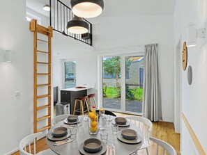 Uma sala de jantar com uma mesa redonda posta para quatro pessoas, uma escada de madeira e vista para o exterior.