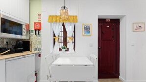 A compact kitchen with a microwave, coffee maker, and a small cabinet. There's a dining table with chairs and a hanging light fixture. A red door and a framed picture are visible on the wall. - GuestReady - Porto's relaxing nook (Porto)