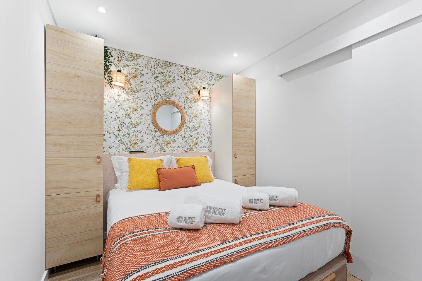 A modern hotel room with a bed, a round mirror, and a floral wallpaper.
