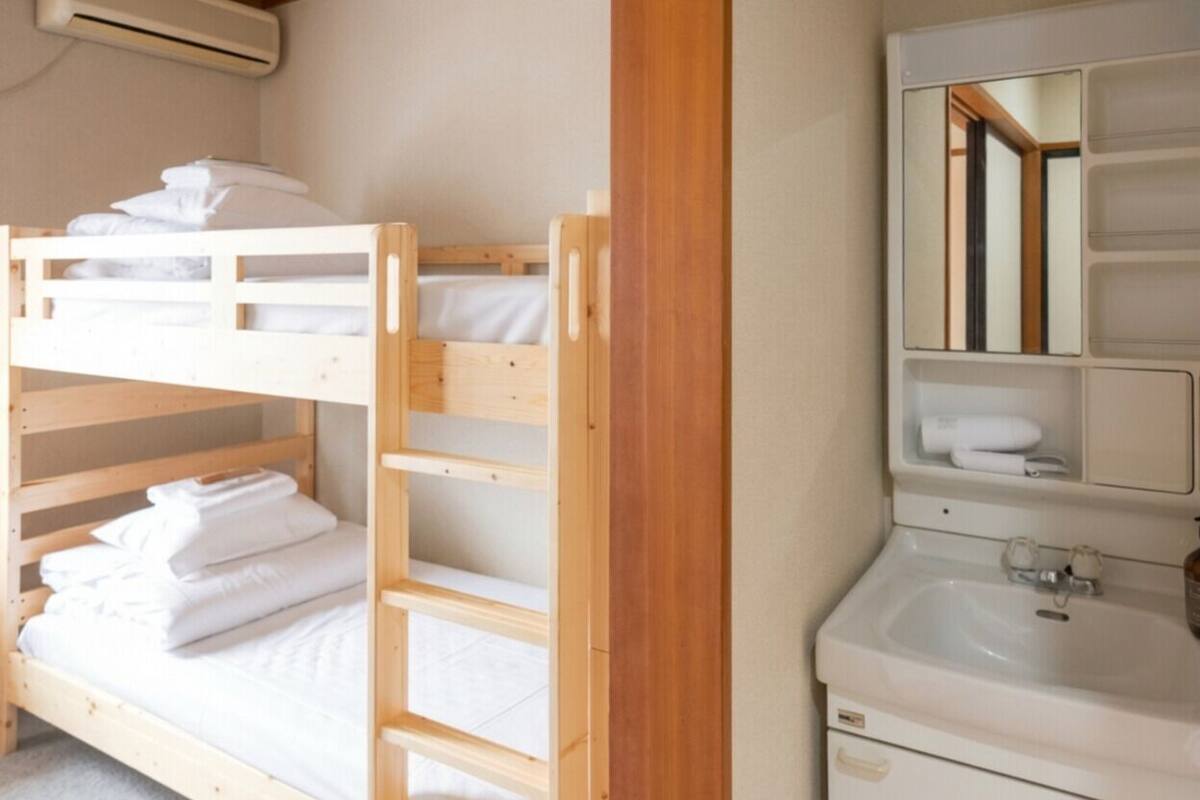 Un lit superposé avec une literie blanche et une échelle en bois, un lavabo blanc avec un miroir au-dessus, et une petite étagère avec des serviettes.