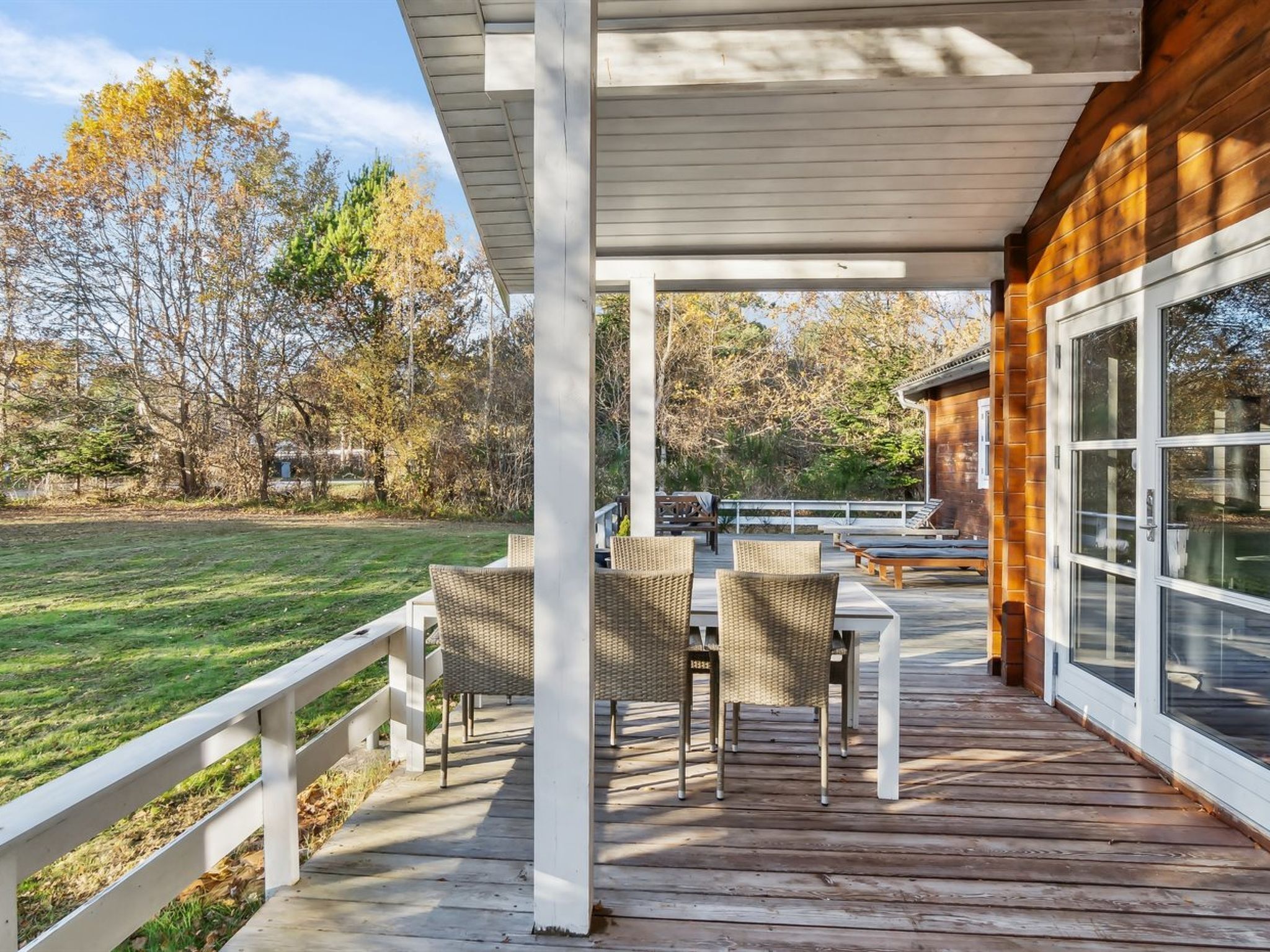 A covered outdoor deck with wicker chairs and a table, overlooking a grassy yard and trees.