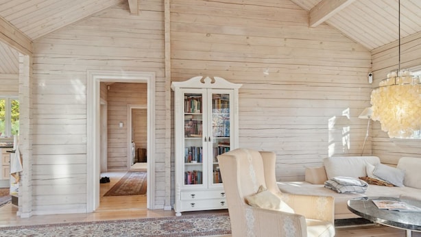 Een ruime kamer met houten wanden, een wit kastje met boeken, een beige fauteuil en een ronde tafel.