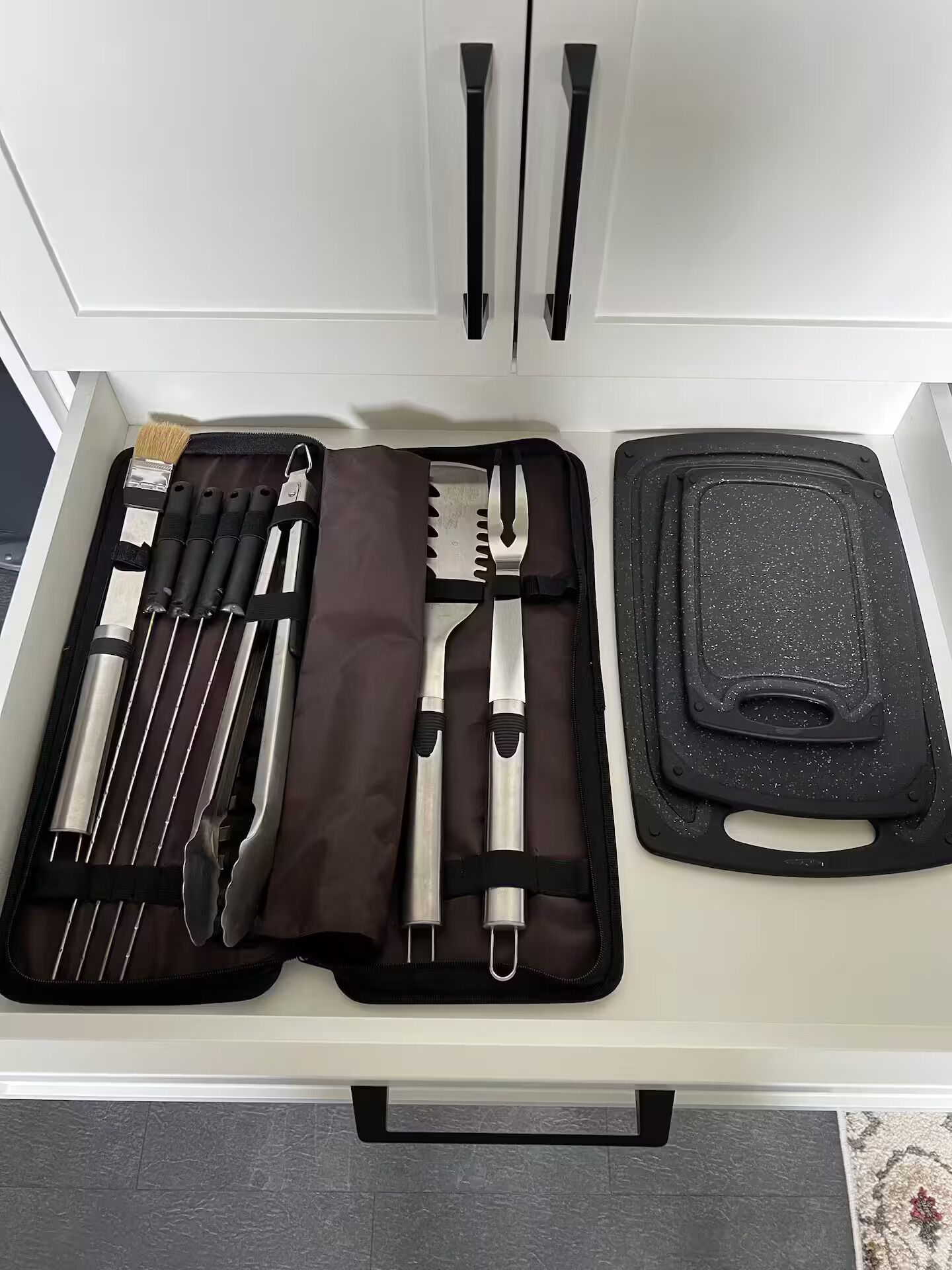 A neatly organized drawer with a set of utensils, a black cutting board, and a brown cloth.