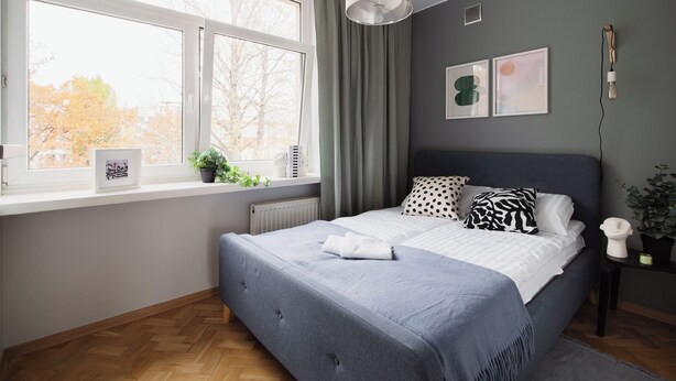 Een slaapkamer met een groot raam, een bed met gestreepte kussens, een radiator en een plant in een pot.