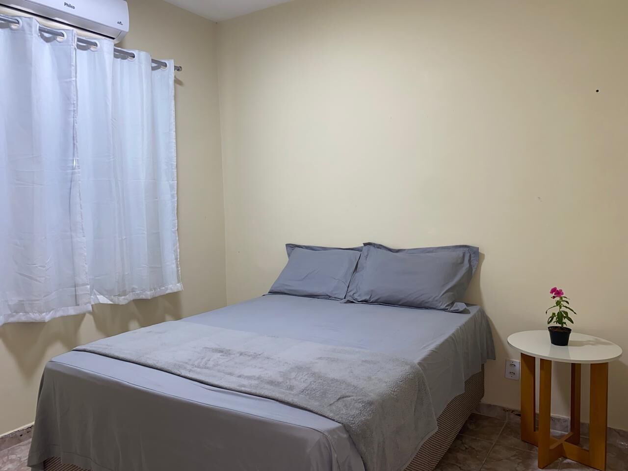 A bedroom with a bed, a small round table with a potted plant, and a window with white curtains.
