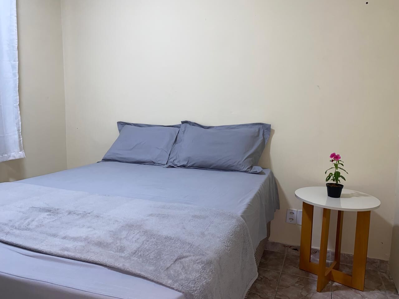 Une chambre à coucher avec un lit, des oreillers, une table de chevet ornée d’une plante en pot et une fenêtre avec un rideau.