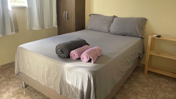 A neatly made bed with a grey bedspread and two rolled towels, a wooden wardrobe, and a small wooden nightstand with a remote control. - Ceo Studio Premium 4 | Comfort and Style close to everything (Aracaju)
