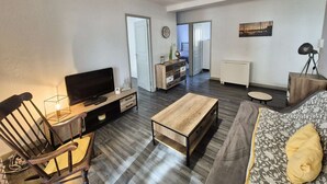 Un salon moderne comprenant un canapé, une table basse, un téléviseur et un meuble en bois. Une porte donne accès à une autre pièce, et une fenêtre offre une vue dégagée.
