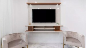 A modern living room with a flat-screen TV mounted on a white wall, two armchairs, and a wooden shelf below the TV. - Spacious 3BR Luxe Escape near NYC (Yonkers)