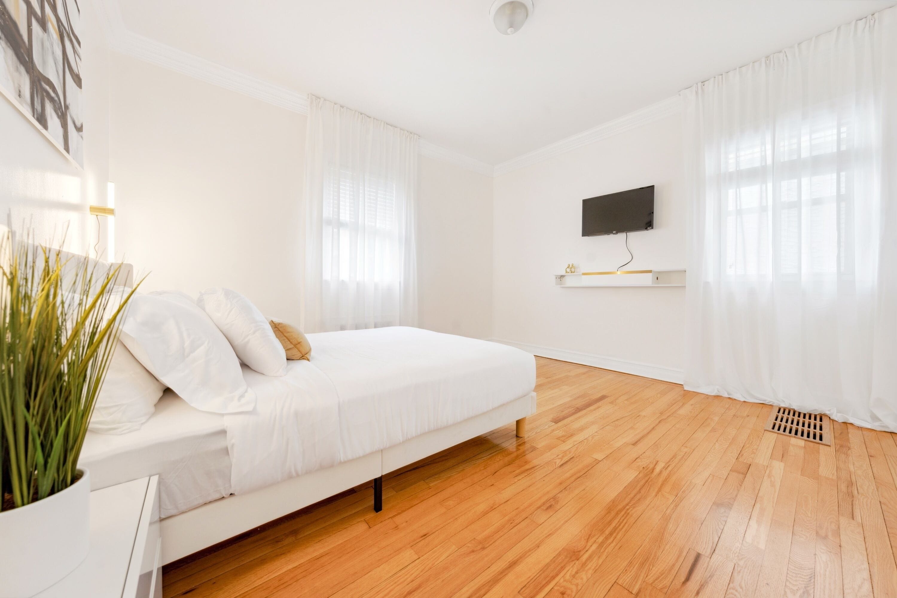 A modern bedroom with a white bed, wooden flooring, a mounted TV, and a potted plant.