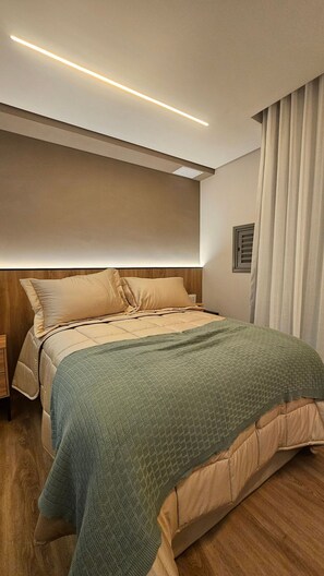 A modern bedroom with a wooden headboard, a large bed with beige bedding, a green knitted throw, and a ceiling light. - Terrae Pinheiros (São Paulo)