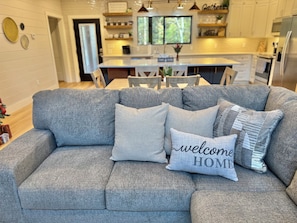 A grey sofa with multiple cushions, including a 'welcome home' pillow, in a modern kitchen with white cabinets and a dining area. - Gracewood Creek - Luxury Cabin, New Heated Pool (Broken Bow)