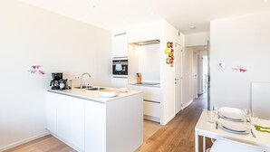 Een moderne keuken met een centraal eiland, een eetgedeelte met een tafel gedekt voor twee personen, en een gang naar de slaapkamers.