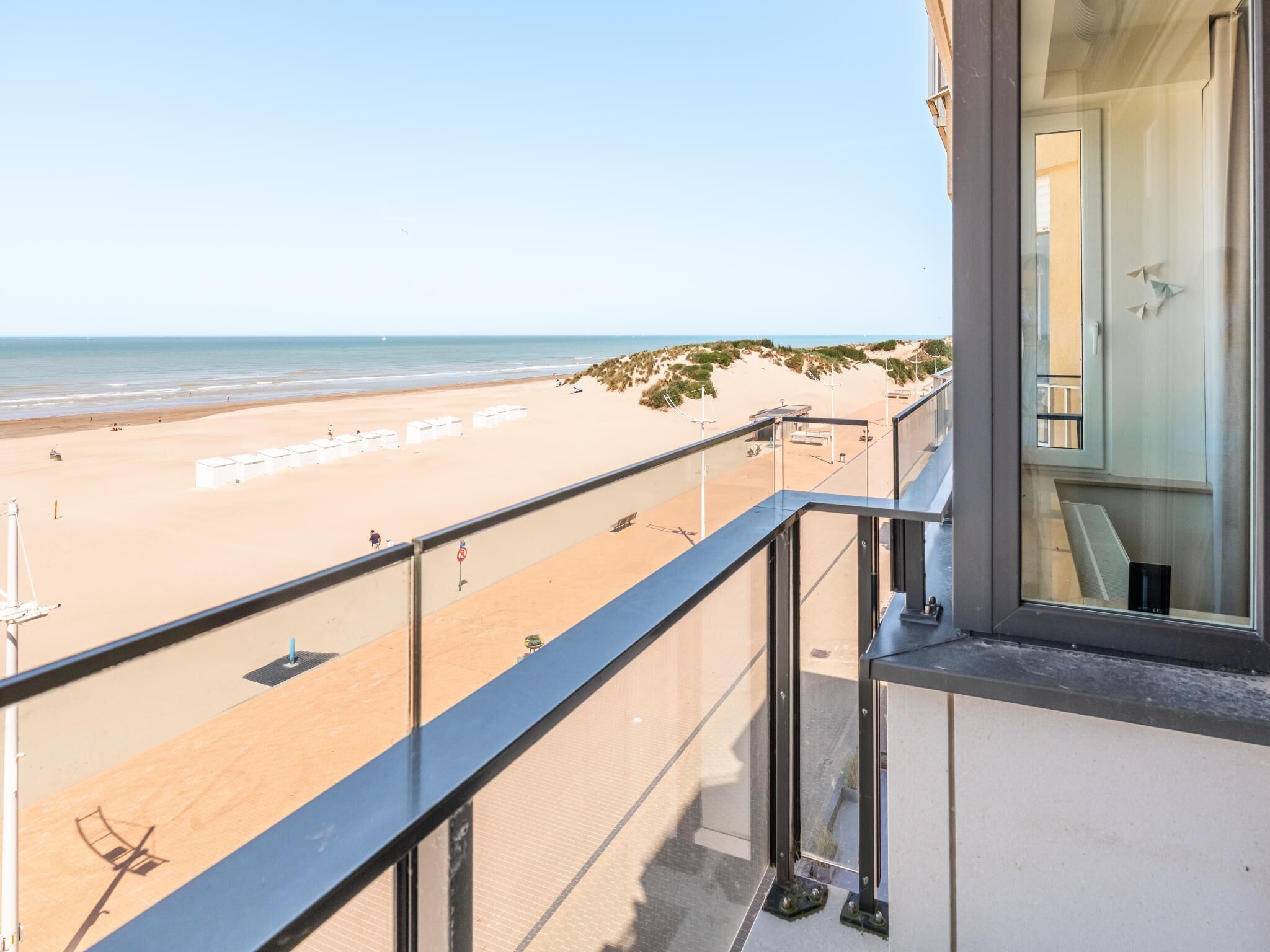 A balcony with a view of a beach, a glass door, and a window reflecting the beach.
