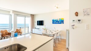 Moderni hotellihuone, jossa on keittiö, ruokailutila ja suuri ikkuna, josta avautuu näkymä merelle.