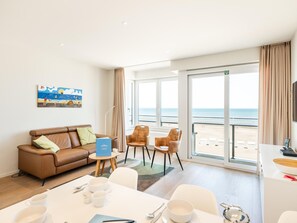 Habitación de hotel con un ventanal grande con vistas a la playa, una zona de comedor con vajilla blanca, un sofá de cuero marrón y un cuadro en la pared.