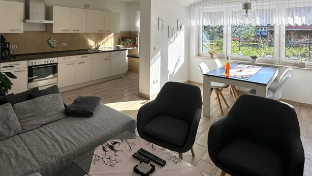 Eine moderne Küche mit weißen Schränken, ein Essbereich mit Tisch und Stühlen sowie ein Wohnzimmer mit Sofa und Couchtisch.