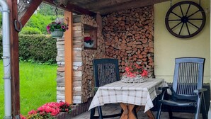 Een overdekt terras met een tafel en stoelen, een stenen muur met een stapel hout en een tuin met bloemen.