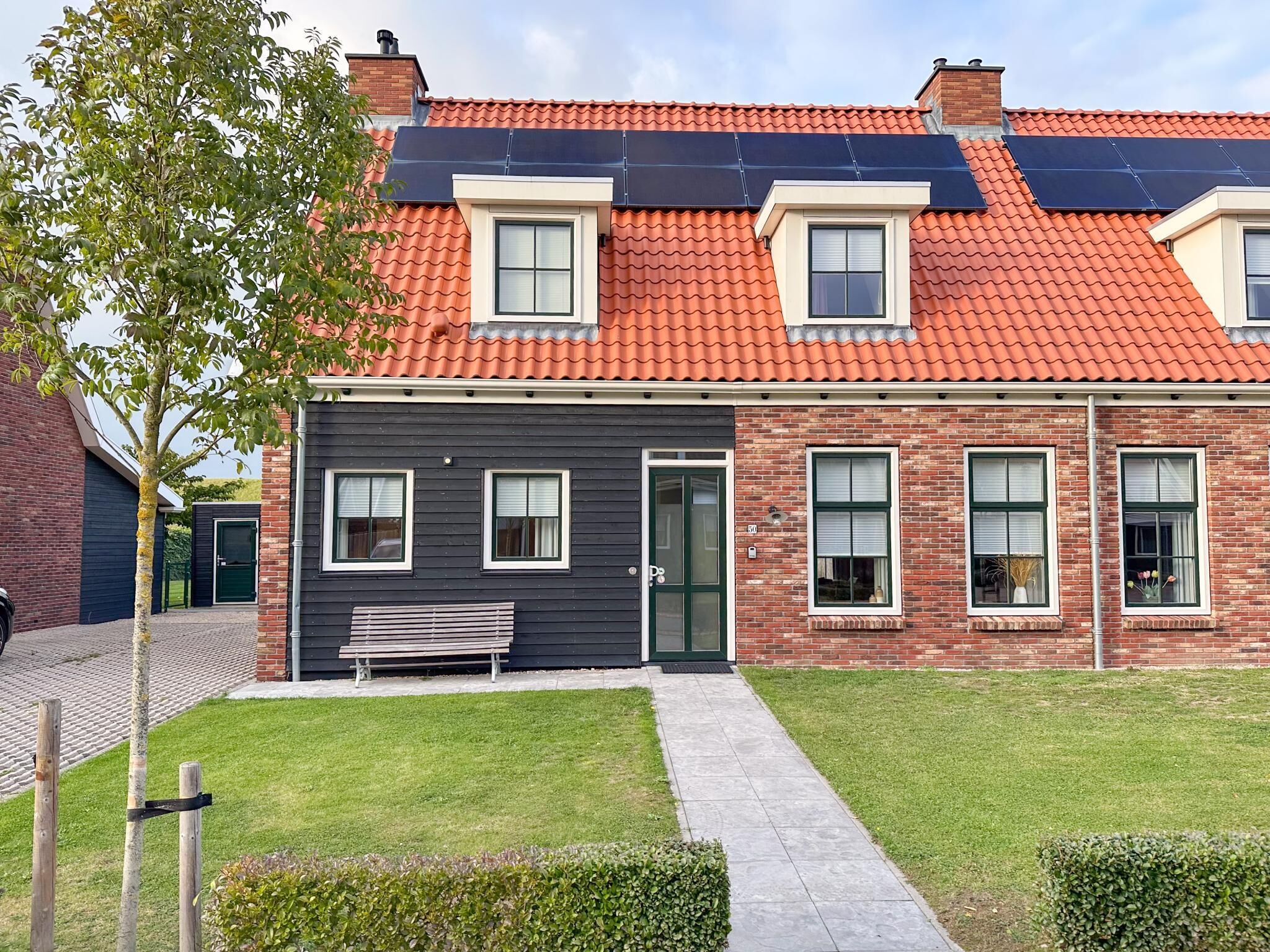 Uma casa moderna de dois andares, com telhado de telhas vermelhas, fachada cinza escura e um pequeno jardim frontal com um banco.