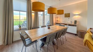 A modern dining area with a wooden table and chairs, large windows with curtains, and pendant lighting. - Wellness villa with sauna, garden and sea nearby (Colijnsplaat)