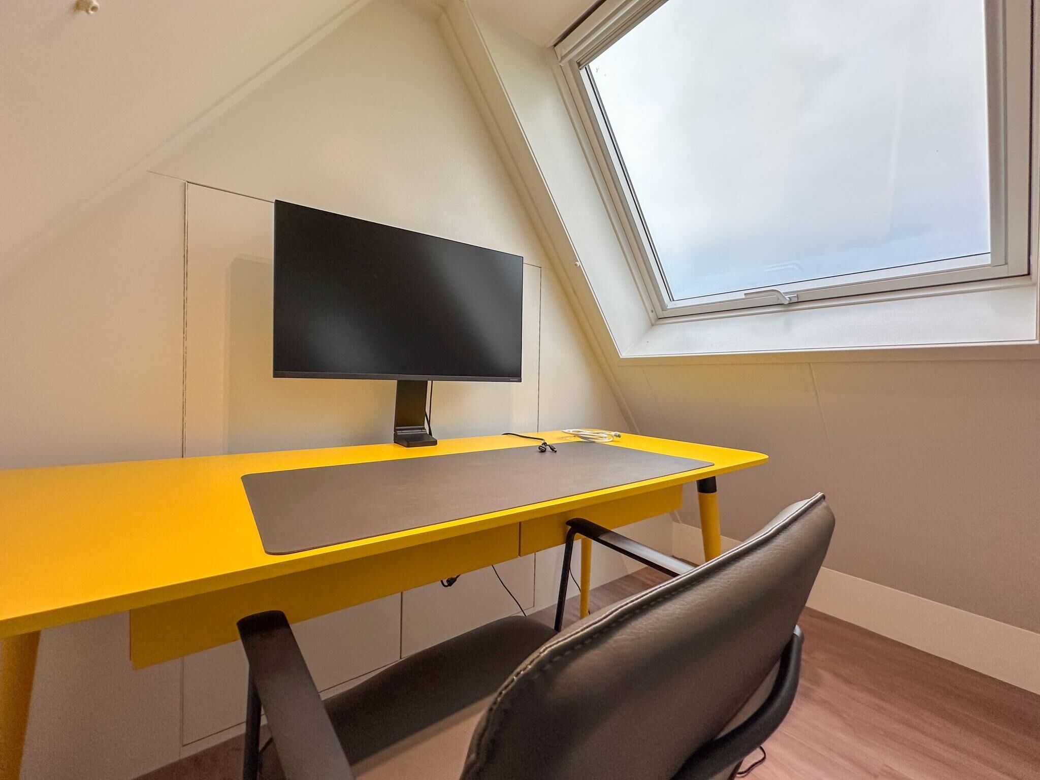 Een kamer met een geel bureau, een zwarte stoel, een flatscreen televisie en een dakraam.
