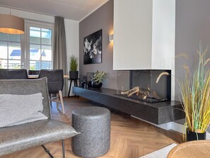 Ein modernes Wohnzimmer mit Kamin, grauem Sofa, Hocker und Blick ins Freie durch große Fenster.