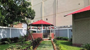 Un sendero en un jardín que conduce a un pabellón con techo rojo, rodeado de abundante vegetación y un muro de ladrillo.