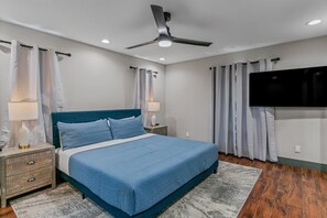 Ein Schlafzimmer mit Bett, Nachttischen, Deckenventilator, Vorhängen, Fernseher und Holzboden.