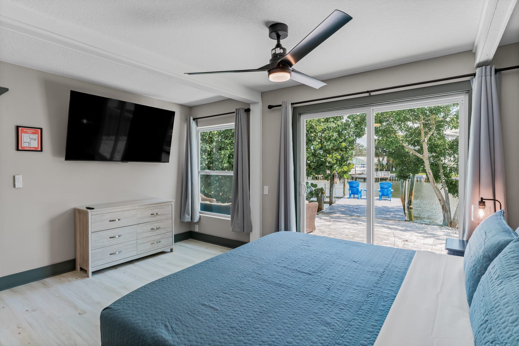A bedroom with a bed, a ceiling fan, a TV, and a view of a water body and greenery outside.