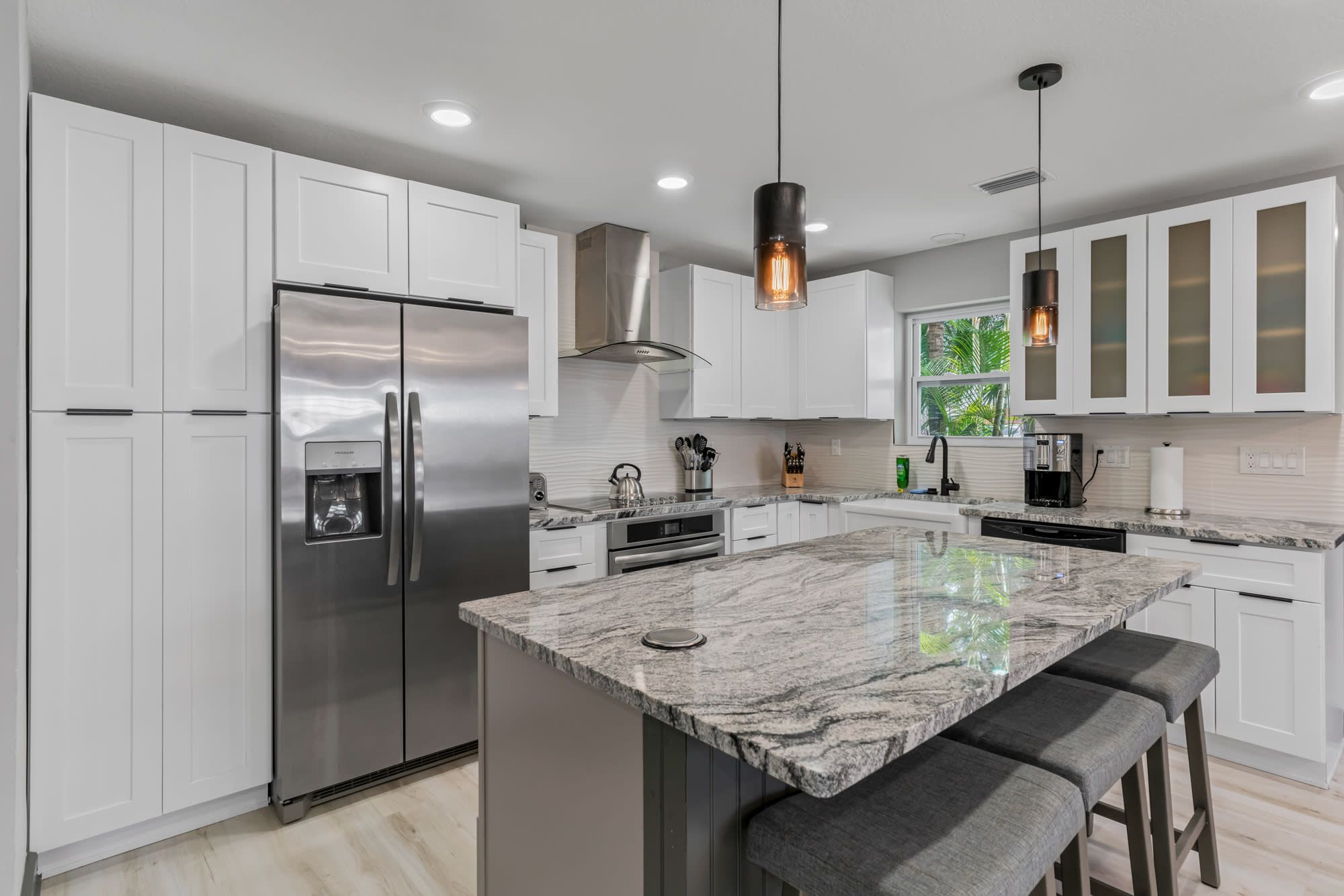 A modern kitchen with white cabinets, a stainless steel refrigerator, a large marble island with bar stools, pendant lighting, and a window with a view of greenery.