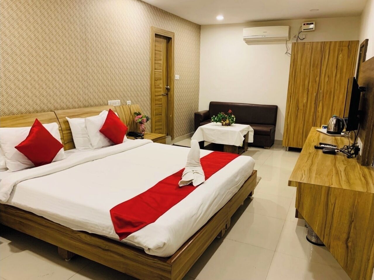 A hotel room with a large bed, a desk, a small table with a vase of flowers, and a wardrobe.