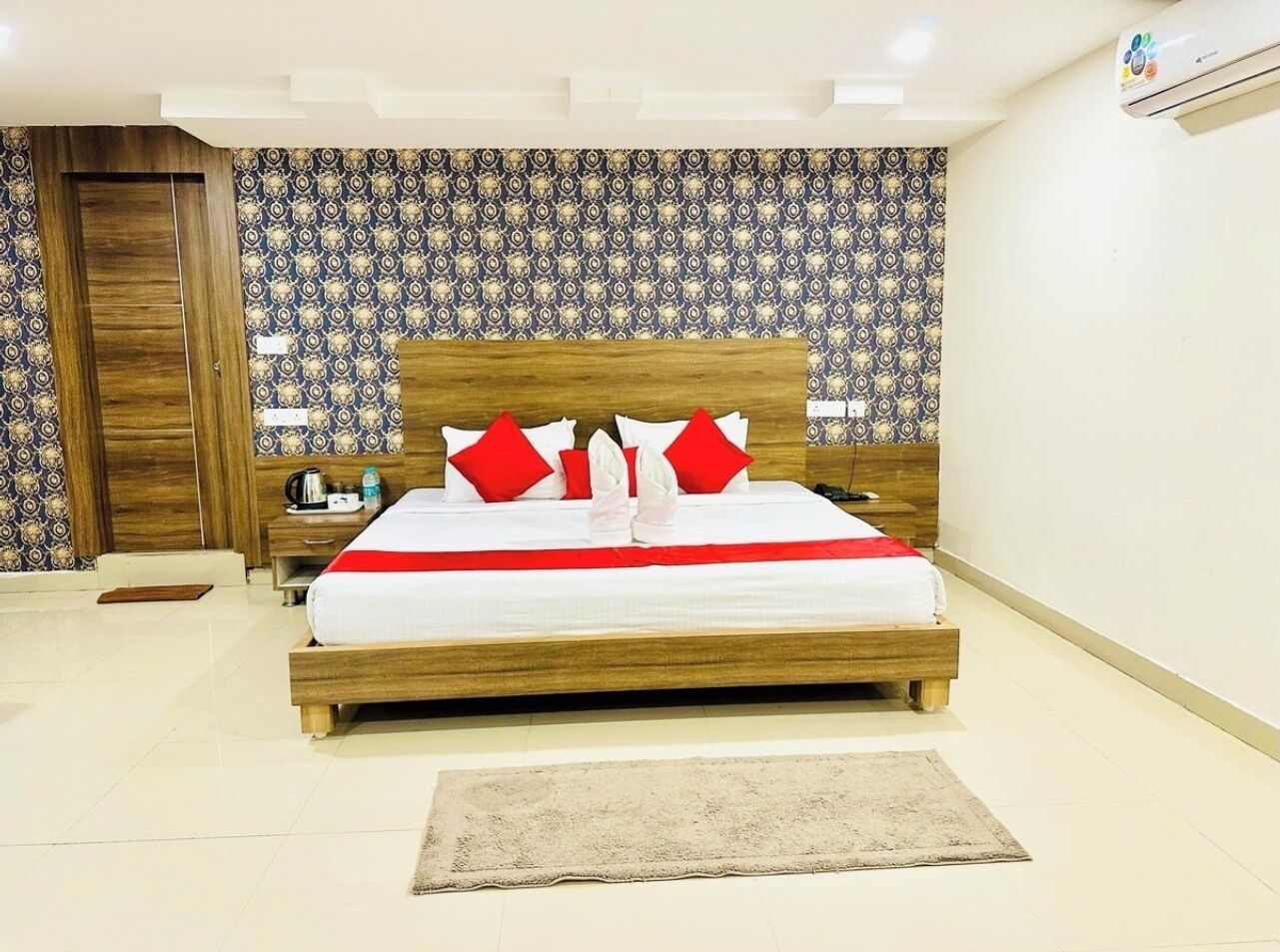 Una cama bien hecha con tablero de madera y almohadas rojas, una mesita de noche con lámpara, y un papel tapiz estampado en la pared.