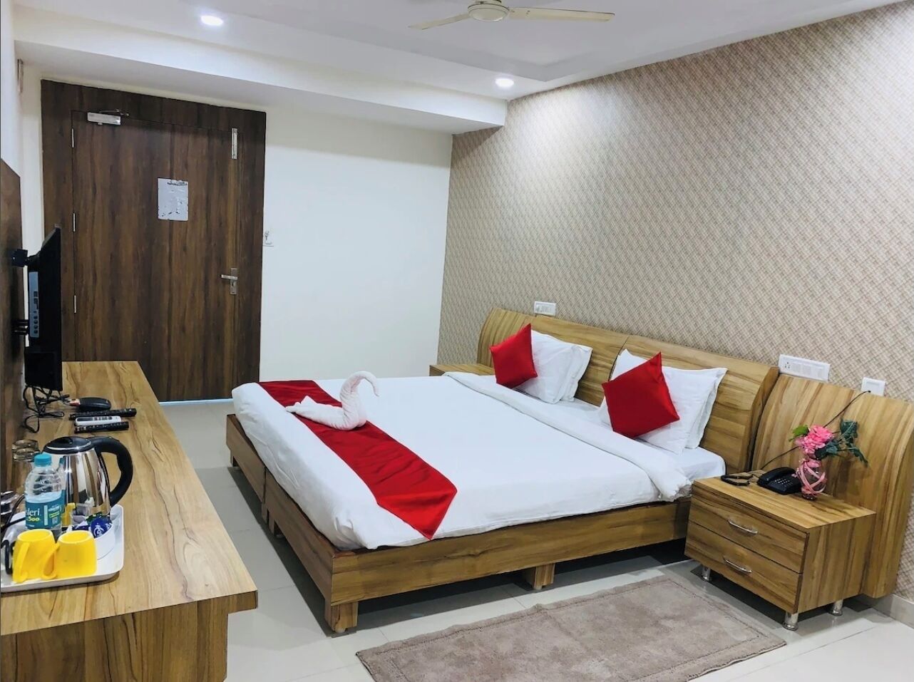 A hotel room with a wooden bed, a desk with a kettle, and a small nightstand with a phone and a vase.