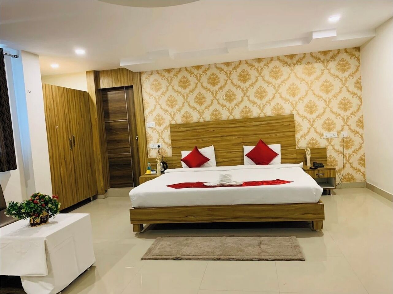 Une chambre d’hôtel avec un lit en bois, des oreillers rouges, une petite table de chevet avec une plante et un papier peint à motifs.