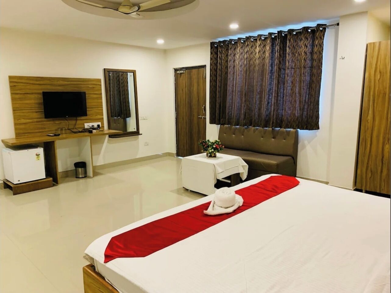 Ein Hotelzimmer mit einem großen Bett, einem Schreibtisch mit Flachbildfernseher, einem kleinen Tisch mit einer Vase, einer Couch und einem großen Fenster mit Vorhängen.