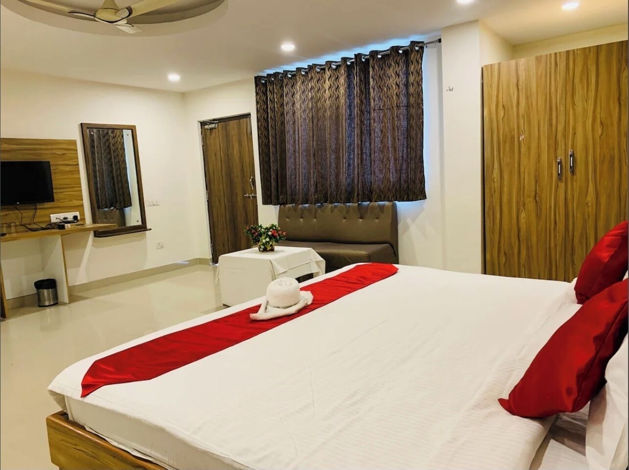 A hotel room with a large bed, a wooden wardrobe, a small table with a vase, a mirror, and a television on a wooden cabinet.