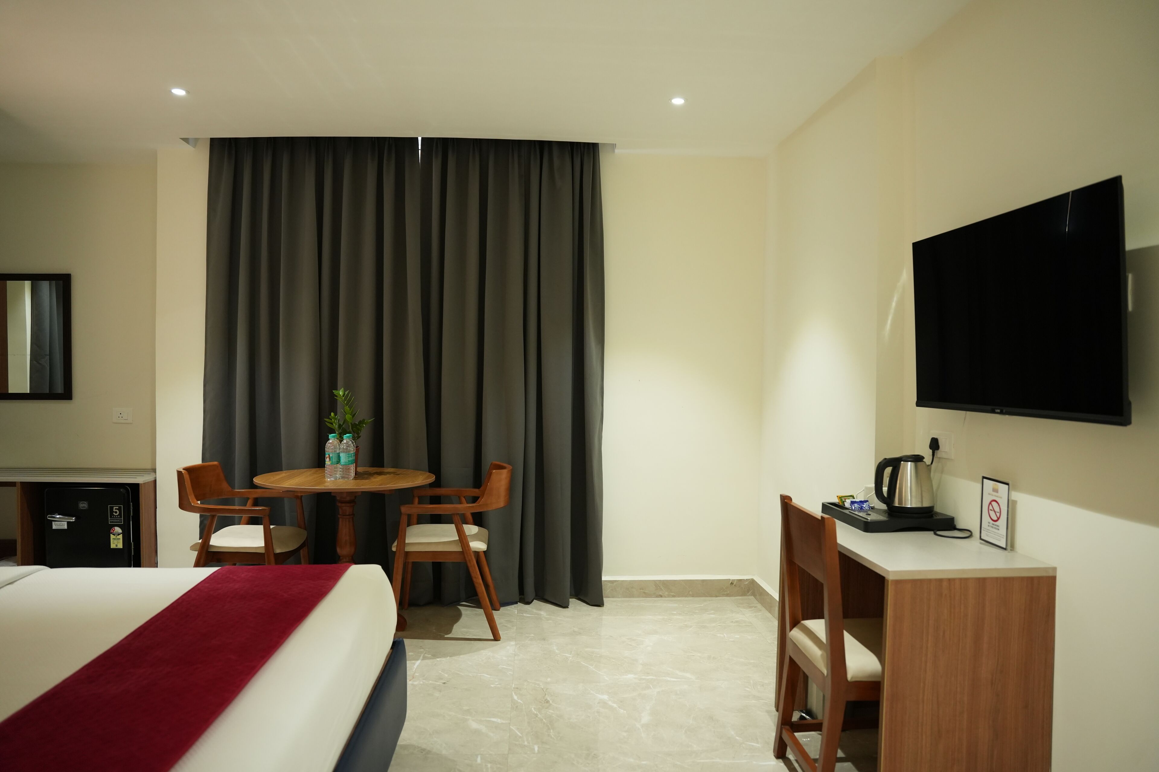 A hotel room with a bed, a desk with a television, a small table with chairs, and a window with curtains.