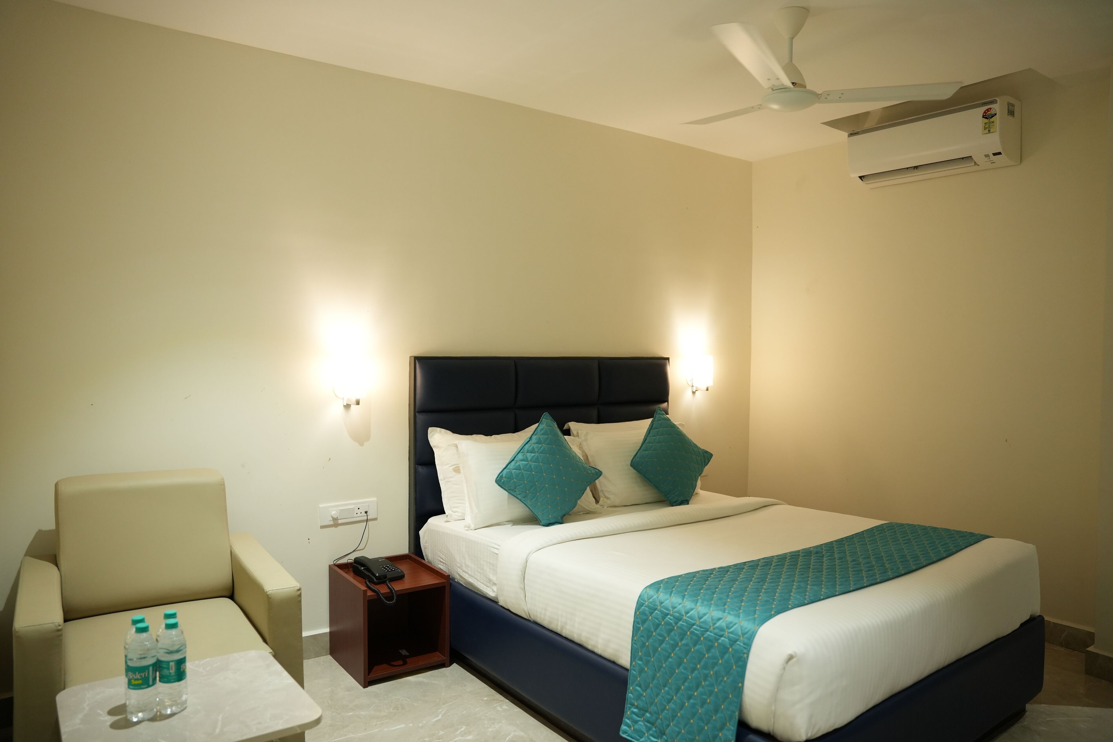 A hotel room with a bed, a chair, a nightstand, and a wall-mounted air conditioner.