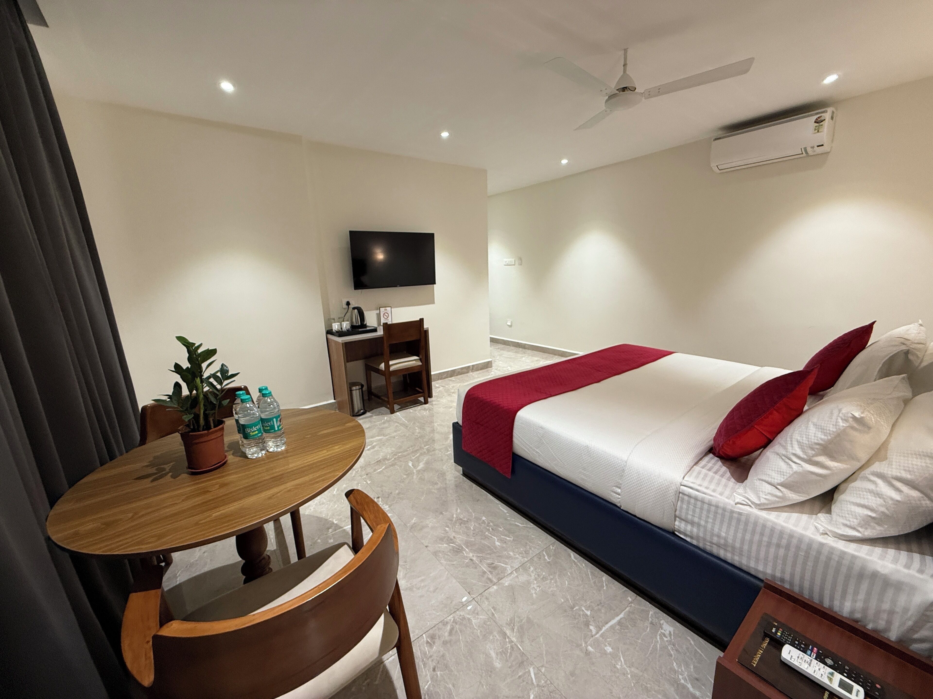 A hotel room with a bed, a desk, a chair, a table with a potted plant and water bottles, a TV, and an air conditioning unit.