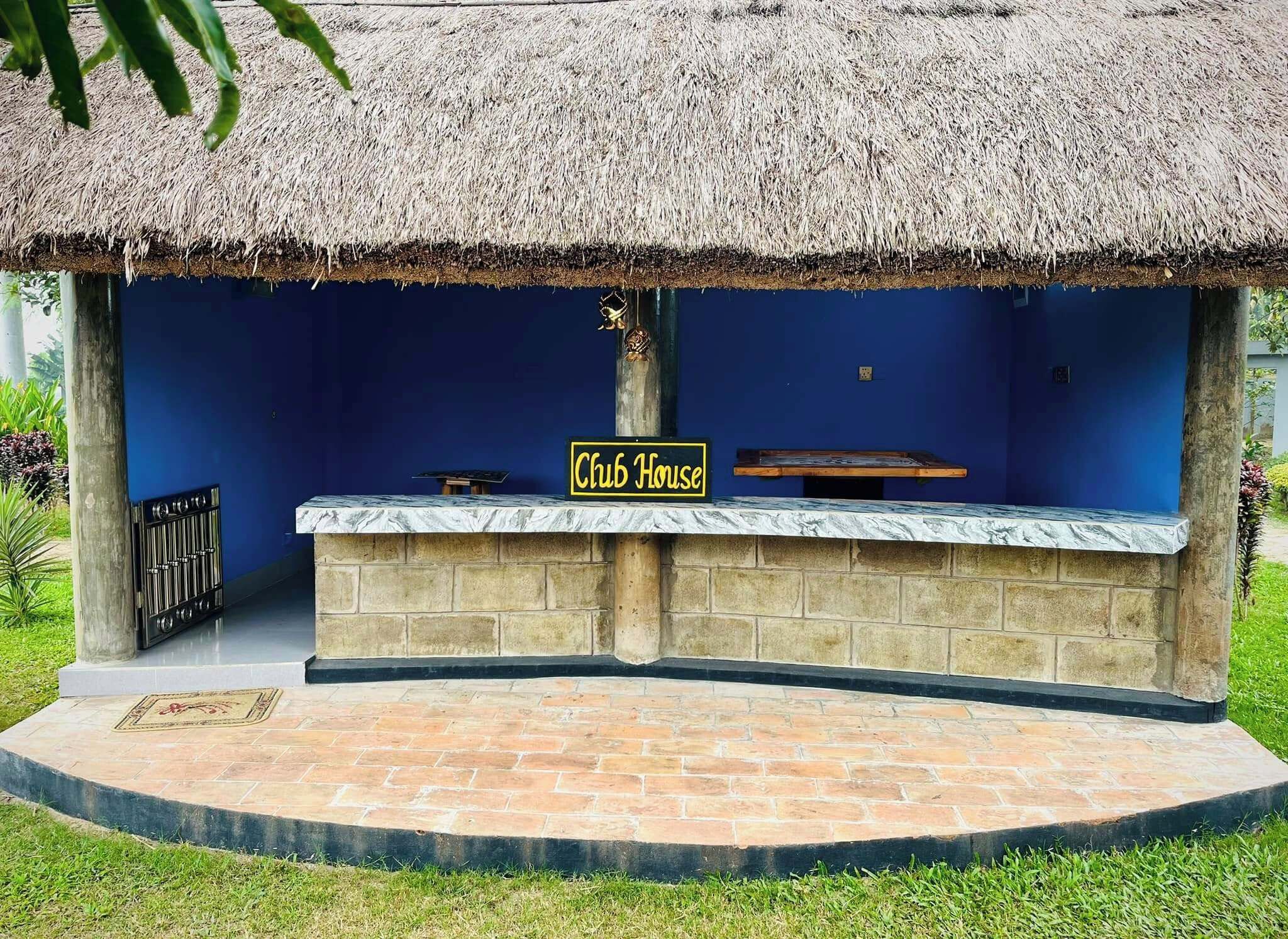 A thatched-roof structure labeled "Club House" with a stone bench and a small table, surrounded by a paved area and greenery.